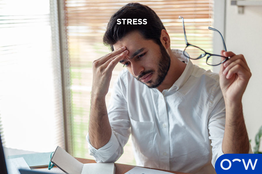 Man with a stressed facial expression, leaning on his hand and holding glasses at a desk.